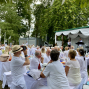 Viele Menschen in weißer Kleidung sitzen bei einer festlichen Veranstaltung im Freien an gedeckten Tischen unter Bäumen. Auf einer kleinen, mit weißen Stoffbahnen dekorierten Bühne treten Musiker vor dem Publikum auf.