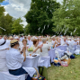Viele Menschen in weißer Kleidung sitzen an langen, weiß gedeckten Tischen bei einem Fest im Park. Zwischen den grünen Bäumen hängen weiße Bänder, während die Gäste applaudieren und gemeinsam essen.