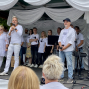 Auf einer mit weißen Stoffbahnen geschmückten Bühne stehen mehrere Menschen in überwiegend weißer Kleidung, während ein Mann mit Mikrofon zum Publikum spricht. Neben ihm warten weitere Sänger und Musiker mit Mikrofonen, Gitarre und Keyboard auf ihren Einsatz.
