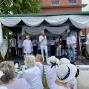 Auf einer dekorierten Freiluftbühne stehen mehrere weiß gekleidete Personen, während ein Mann mit Mikrofon zum Publikum spricht. Vor der Bühne sitzen zahlreiche Zuschauerinnen und Zuschauer in heller Kleidung und weißen Hüten an Tischen.