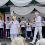 Auf einer kleinen Bühne spricht eine Frau in weißer Kleidung mit einem Mikrofon vor einem Publikum. Hinter ihr stehen mehrere Personen in weißen „über LEBEN“-T-Shirts, während ein Mann applaudiert und rechts ein Gitarrist zu sehen ist.