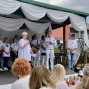 Eine Frau spricht mit einem Mikrofon auf einer weiß geschmückten Bühne, während mehrere Personen in weißen Shirts hinter ihr stehen. Vor der Bühne sitzt Publikum, und Musikinstrumente sowie Mikrofonständer deuten auf eine Veranstaltung oder Aufführung im Freien hin.