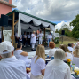 Eine Gruppe weiß gekleideter Menschen sitzt bei einer Veranstaltung im Freien vor einer kleinen geschmückten Bühne. Auf der Bühne spricht eine Frau ins Mikrofon, während weitere Personen und Musikausrüstung im Hintergrund zu sehen sind.