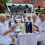Eine Gruppe älterer Menschen in weißer Kleidung sitzt bei einer festlich gedeckten Tafel im Freien und lächelt in die Kamera. Im Hintergrund sind weitere Gäste, eine kleine Bühne mit weißen Vorhängen und ein rotes Gebäude zu sehen.