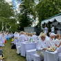 Viele Menschen in weißer Kleidung sitzen im Freien an weiß gedeckten Tischen in einem grünen Park. Rechts befindet sich eine dekorierte Bühne, auf der zwei Männer vor dem Publikum sprechen.