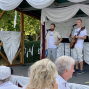Auf einer mit weißen Stoffbahnen dekorierten Außenbühne stehen zwei Männer vor Mikrofonen, einer davon mit einer Gitarre. Im Vordergrund sitzen Zuschauer an Tischen, während im Hintergrund grüne Bäume und Hecken zu sehen sind.
