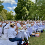 Eine große Gruppe festlich in Weiß gekleideter Menschen sitzt an gedeckten Tischen in einem grünen Park und applaudiert. Über ihnen hängen weiße Bänder in den Bäumen, während bei sonnigem Wetter eine gesellige Feier im Freien stattfindet.