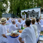 Viele weiß gekleidete Menschen sitzen im Freien an langen Tischen und hören einer Person auf einer geschmückten Bühne zu. Die Veranstaltung findet unter Bäumen statt, mit weißen Stoffdekorationen, Lautsprechern und gedeckten Tischen voller Speisen und Getränke.
