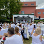 Viele Menschen in weißer Kleidung sitzen im Freien an langen gedeckten Tischen und schauen auf eine kleine Bühne mit Musikern. Im Hintergrund stehen ein rotes Gebäude, große Bäume mit weißen Bändern und ein bewölkter Himmel.