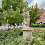 Eine steinerne Frauenstatue mit Blumen steht in einem üppigen Garten zwischen Rosen und grünen Bäumen. Im Hintergrund sitzen viele weiß gekleidete Menschen bei einer Veranstaltung vor einem roten Gebäude unter bewölktem Himmel.