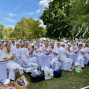 Viele Menschen in weißer Kleidung sitzen an langen, weiß gedeckten Tischen bei einem Fest im Park. Zwischen großen grünen Bäumen hängen weiße Bänder, während die Gäste essen, trinken und applaudieren.