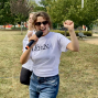 Eine fröhliche Frau mit Sonnenbrille hält in einem Park ein Mikrofon und hebt begeistert die Hand. Hinter ihr steht eine weitere Person, während im Hintergrund Bäume, Wege und Parkbänke zu sehen sind.