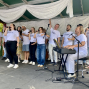 Eine Gruppe von Sängerinnen und Sängern in weißen T-Shirts tritt auf einer kleinen Bühne auf und singt mit Mikrofonen. Rechts begleitet ein Mann am Keyboard die Gruppe, während weiße Stoffbahnen die Bühne schmücken.