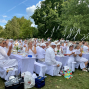 Eine große Gruppe überwiegend weiß gekleideter Menschen sitzt an festlich gedeckten Tischen im Grünen und applaudiert. Über ihnen hängen weiße Bänder in den Bäumen, während der Himmel blau und sonnig ist.