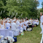 Eine große Gruppe überwiegend weiß gekleideter Menschen sitzt im Freien an gedeckten Tischen unter Bäumen. Rechts steht ein Mann mit Mikrofon neben einer kleinen Bühne und spricht zum Publikum.