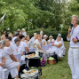 Ein Mann in weißer Kleidung spricht mit einem Mikrofon zu einer Gruppe von Menschen, die an weiß gedeckten Tischen im Freien sitzen. Die Veranstaltung findet in einem grünen Park statt, und viele Gäste tragen ebenfalls weiße Kleidung.