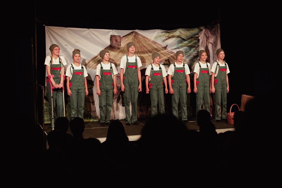 Mehrere Kinder stehen in identischen grünen Latzhosen, weißen T-Shirts und grauen Mützen auf einer Theaterbühne vor einem bemalten Bühnenbild mit Hausdach. Im Vordergrund sind die Silhouetten des Publikums zu sehen, während rechts ein Korb und links eine Krücke als Requisiten erkennbar sind.
