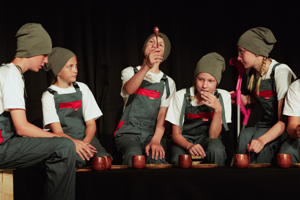 Mehrere Kinder in grauen Latzhosen, weißen T-Shirts und Mützen sitzen auf einer Bühne vor einem dunklen Vorhang. Vor ihnen stehen kleine Holzbecher, während ein Kind einen Holzlöffel hochhält und die anderen aufmerksam zuschauen.