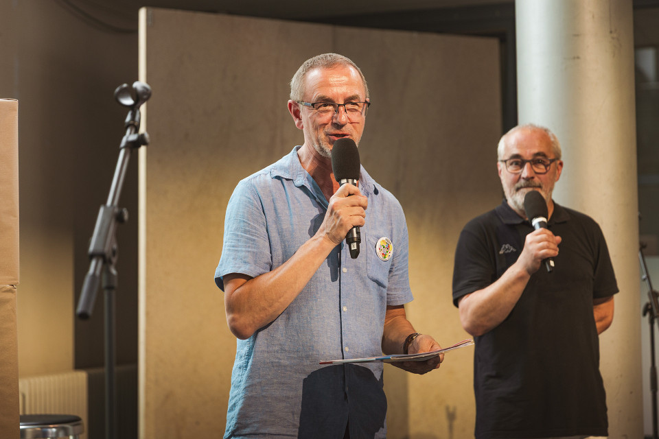 Zwei Männer stehen in einem Innenraum vor einer Betonwand und sprechen jeweils in ein Mikrofon. Der Mann im Vordergrund trägt ein hellblaues Hemd und hält Unterlagen in der Hand, während im Hintergrund ein weiterer Mann in Schwarz zu sehen ist.