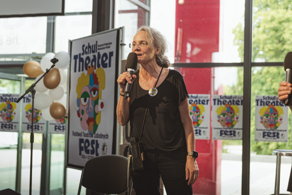 Eine Frau steht bei einer Veranstaltung in einem hellen Foyer und spricht mit einem Mikrofon vor Plakaten eines Schultheater-Festivals. Neben ihr sind Luftballons, ein Mikrofonständer und große Fenster mit Blick ins Grüne zu sehen.
