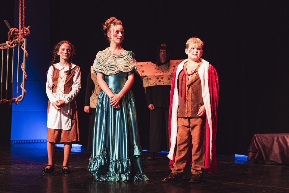 Drei Kinder stehen in fantasievollen Kostümen auf einer dunkel beleuchteten Theaterbühne. Links sind Seile und Bühnenelemente zu sehen, während im Hintergrund weitere verkleidete Personen im Schatten stehen.