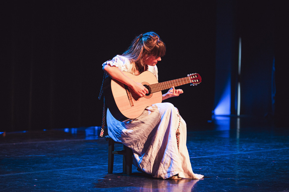Eine junge Frau sitzt auf einem Hocker auf einer dunklen Bühne und spielt konzentriert eine klassische Gitarre. Blaues und warmes Bühnenlicht beleuchten sie und ihr langes helles Kleid vor einem fast schwarzen Hintergrund.