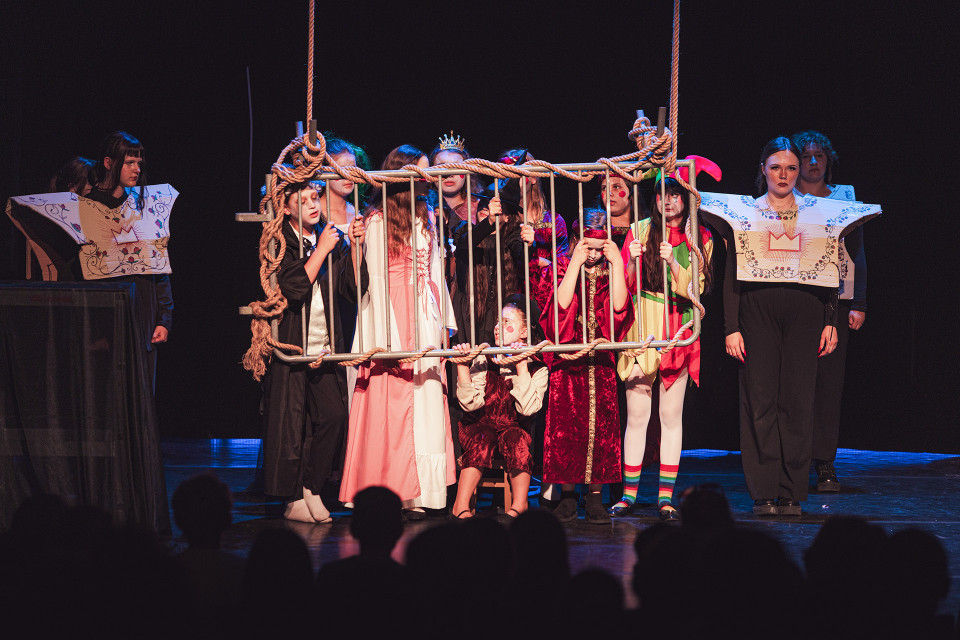 Auf einer Theaterbühne stehen mehrere Kinder und Jugendliche in fantasievollen Kostümen um einen hängenden Käfig mit Seilen. Das dunkle Bühnenlicht und die sichtbaren Zuschauer im Vordergrund lassen die Szene wie eine dramatische Aufführung wirken.