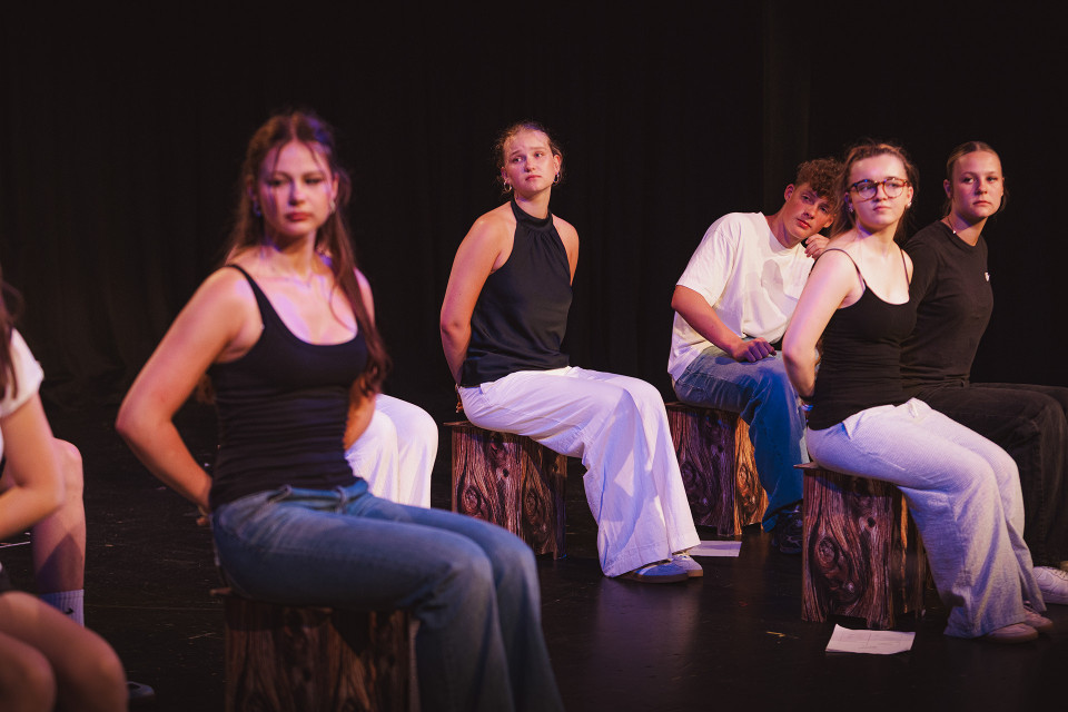 Mehrere Jugendliche sitzen auf einer dunklen Bühne auf hölzernen Hockern und blicken ernst in verschiedene Richtungen. Die Szene ist warm und farbig beleuchtet, während der schwarze Hintergrund den Eindruck einer Theaterprobe oder Aufführung verstärkt.
