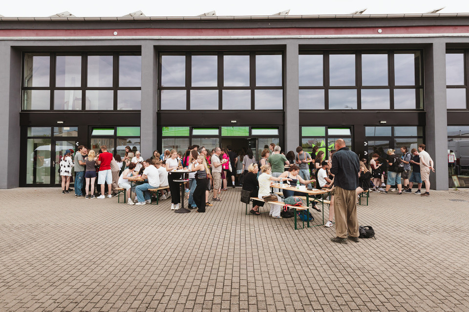 Vor einem modernen Gebäude mit großen Fenstern versammeln sich viele Menschen auf einem gepflasterten Platz. Einige sitzen an Biertischen und essen oder unterhalten sich, während andere in Gruppen vor den Eingängen stehen.