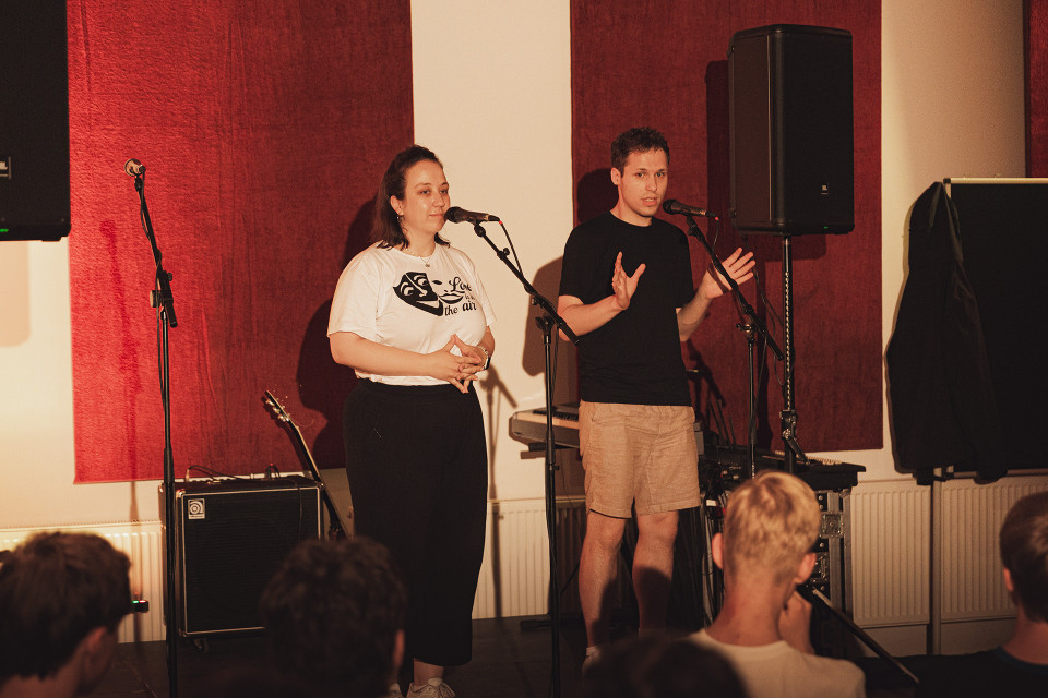 Zwei Personen stehen auf einer kleinen Bühne vor Mikrofonen und sprechen zu einem Publikum. Im Hintergrund sind rote Wandpaneele, Lautsprecher und Musikequipment zu sehen.
