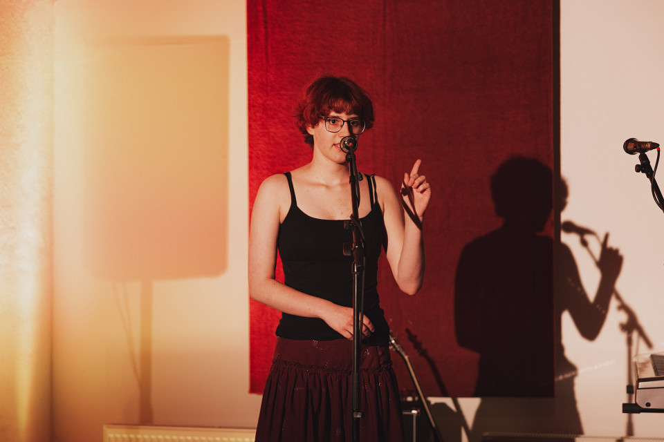 Eine Frau mit Brille steht auf einer Bühne vor einem Mikrofon und hebt einen Finger, als würde sie sprechen oder singen. Hinter ihr wirft das warme Licht einen deutlichen Schatten auf die Wand mit rotem Hintergrund.