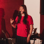 Eine junge Frau in einem roten T-Shirt steht auf einer Bühne vor einem Mikrofon und spricht oder singt. Im Hintergrund sind rote Vorhänge, Musikinstrumente und ein großer Lautsprecher zu sehen.