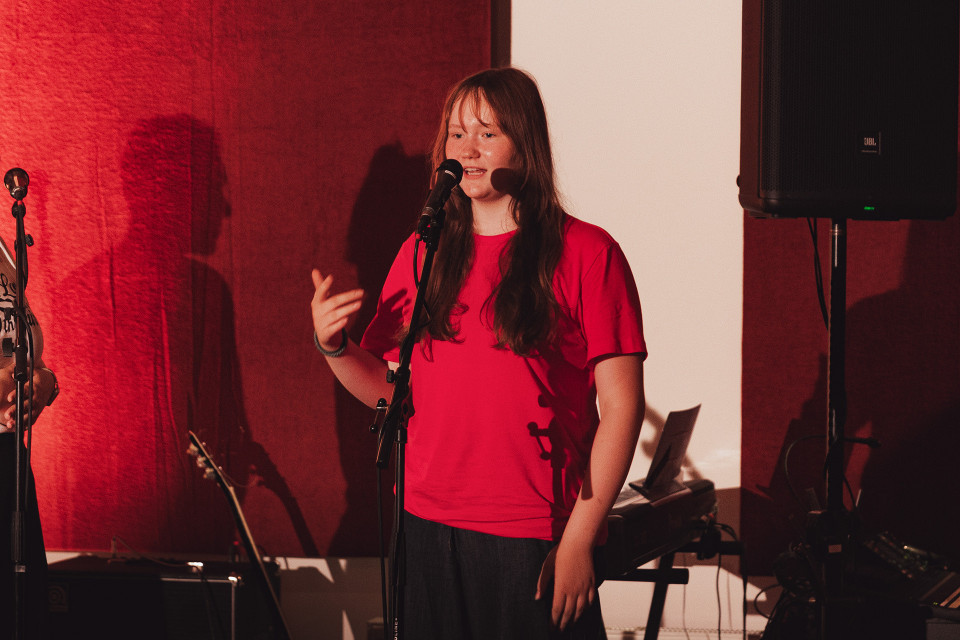 Eine junge Frau in einem roten T-Shirt steht auf einer Bühne vor einem Mikrofon und spricht oder singt. Im Hintergrund sind rote Vorhänge, Musikinstrumente und ein großer Lautsprecher zu sehen.