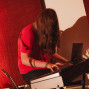 Eine Person mit langen Haaren und rotem T-Shirt sitzt an einem Digitalpiano und spielt darauf. Der Raum ist warm beleuchtet, mit roten Vorhängen im Hintergrund und einem Gitarrenhals am linken Bildrand.