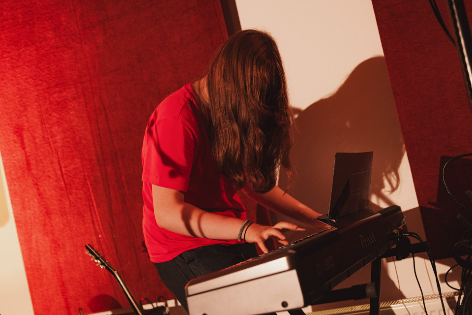 Eine Person mit langen Haaren und rotem T-Shirt sitzt an einem Digitalpiano und spielt darauf. Der Raum ist warm beleuchtet, mit roten Vorhängen im Hintergrund und einem Gitarrenhals am linken Bildrand.