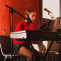 Eine junge Frau sitzt in einem Musikstudio an einem E-Piano und singt in ein Mikrofon. Im Hintergrund sind rote Akustikpaneele, weitere Mikrofonständer und Musikequipment zu sehen.