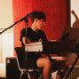 Ein junger Musiker sitzt an einem elektronischen Keyboard und schaut konzentriert auf die Noten oder ein Display. Um ihn herum stehen Mikrofone und Kabel, während warmes Licht vor einem roten Vorhang eine intime Studioatmosphäre schafft.
