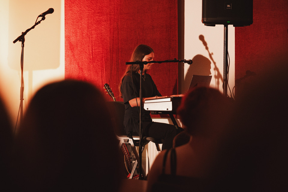 Eine Musikerin sitzt auf einer kleinen Bühne am Keyboard, umgeben von Mikrofonen und warmem, rötlichem Licht. Im Vordergrund sind die dunklen Silhouetten des Publikums zu sehen, während ein Lautsprecher und Schatten an der Wand die Konzertatmosphäre verstärken.