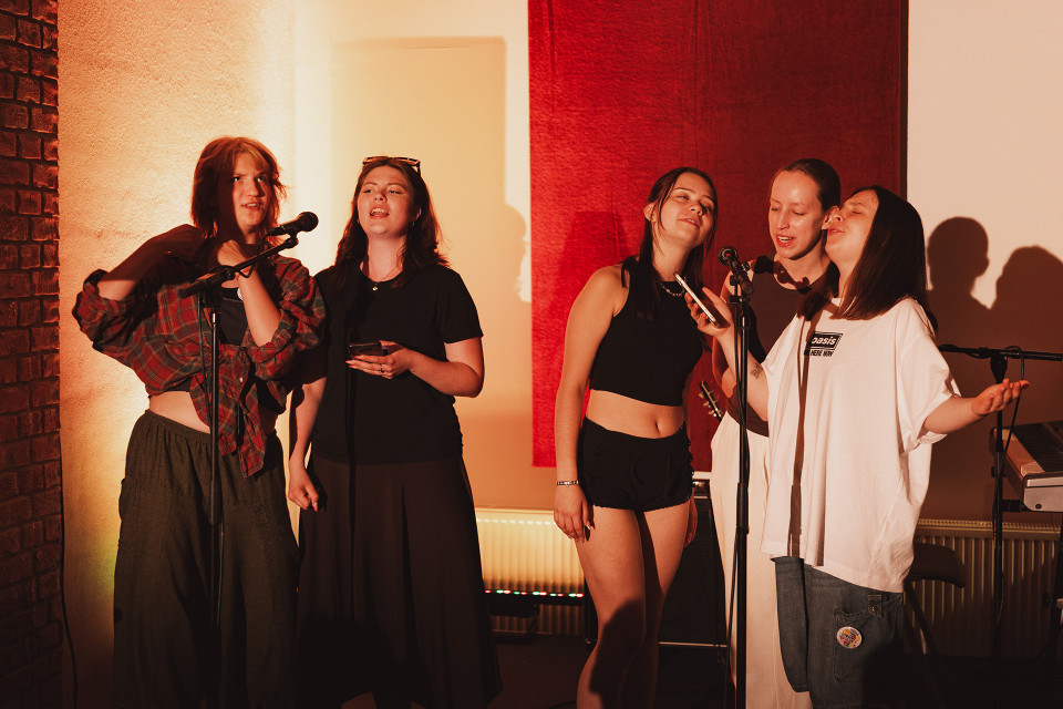 Fünf junge Frauen stehen in warmem Bühnenlicht vor Mikrofonen und singen gemeinsam. Im Hintergrund sind eine rote Wandverkleidung, Schatten an der Wand und ein Keyboard am rechten Rand zu sehen.
