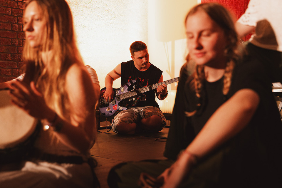In einem warm beleuchteten Raum sitzt ein Mann auf dem Boden und spielt Bassgitarre, während um ihn herum mehrere Personen entspannt zuhören oder musizieren. Der Vordergrund ist unscharf und zeigt junge Menschen in lockerer Atmosphäre, wodurch der Fokus auf dem Musiker in der Mitte liegt.