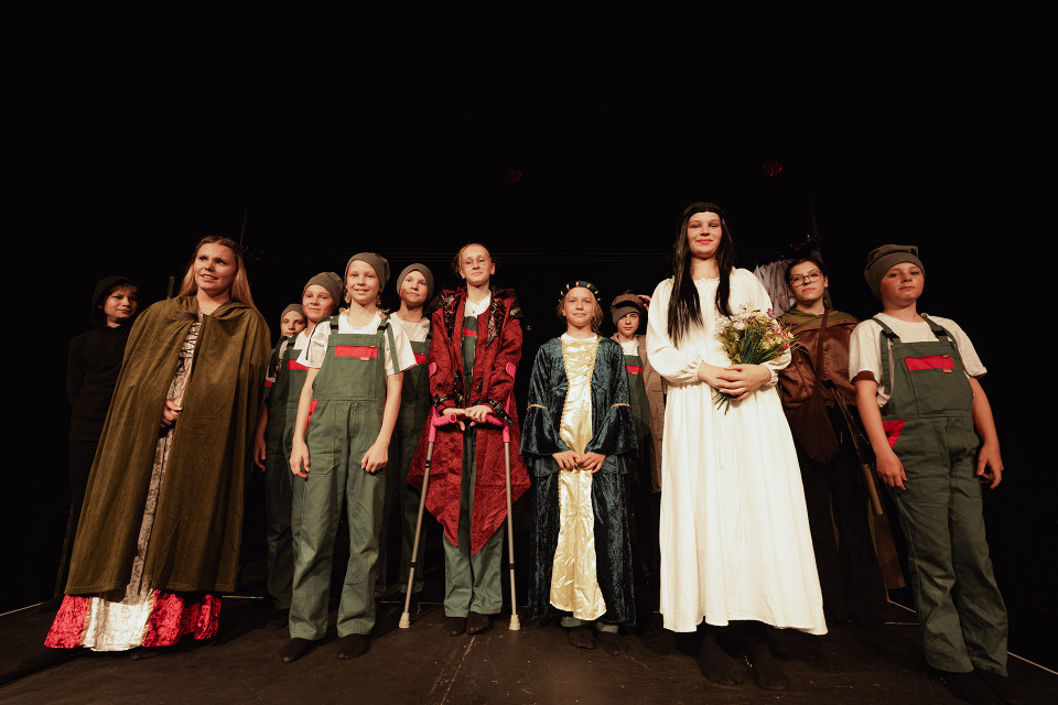Eine Gruppe von Kindern steht auf einer dunklen Theaterbühne in verschiedenen Kostümen, darunter mittelalterliche Gewänder, Overalls und Mützen. Im Vordergrund hält ein Mädchen in einem weißen Kleid einen Blumenstrauß, während die anderen wie nach einer Aufführung posieren.