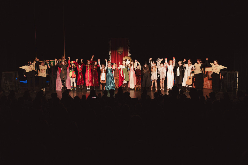 Eine Gruppe von Schauspielerinnen und Schauspielern in bunten Kostümen steht auf einer Theaterbühne und hält sich zum Schlussapplaus an den Händen. Im Vordergrund sind die dunklen Silhouetten des Publikums zu sehen, während die Bühne warm beleuchtet ist.