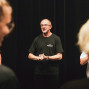Ein Mann in einem schwarzen T-Shirt steht vor einem dunklen Vorhang und spricht zu einer Gruppe von Menschen. Im Vordergrund sind unscharf zwei Personen zu sehen, die den Blick auf die Szene wie einen Rahmen einfassen.