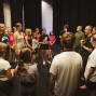 Eine Gruppe von Jugendlichen und Erwachsenen steht im Kreis auf einer Bühne vor schwarzen Vorhängen. Ein Mann spricht zur Gruppe, die aufmerksam zuhört, als ob sie an einer Theaterprobe oder einem Workshop teilnimmt.