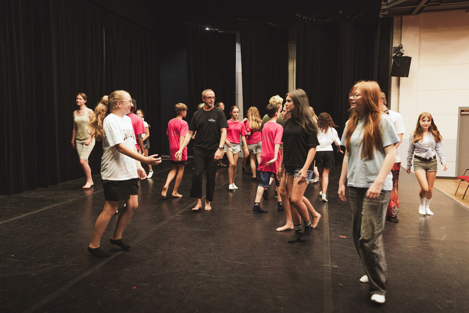 Auf einer Theaterbühne bewegen sich viele Jugendliche und ein erwachsener Mann zwischen schwarzen Vorhängen, offenbar bei einer Probe oder einem Workshop. Einige tragen pinke Shirts, andere Freizeitkleidung, und die Szene wirkt lebendig und ungezwungen.