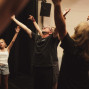Eine Gruppe von Menschen steht in einem Raum, vermutlich bei einer Theater- oder Bewegungsprobe, und streckt die Arme nach oben. In der Mitte leitet ein Mann mit Brille die Übung, während die Teilnehmenden seine Bewegungen nachahmen.