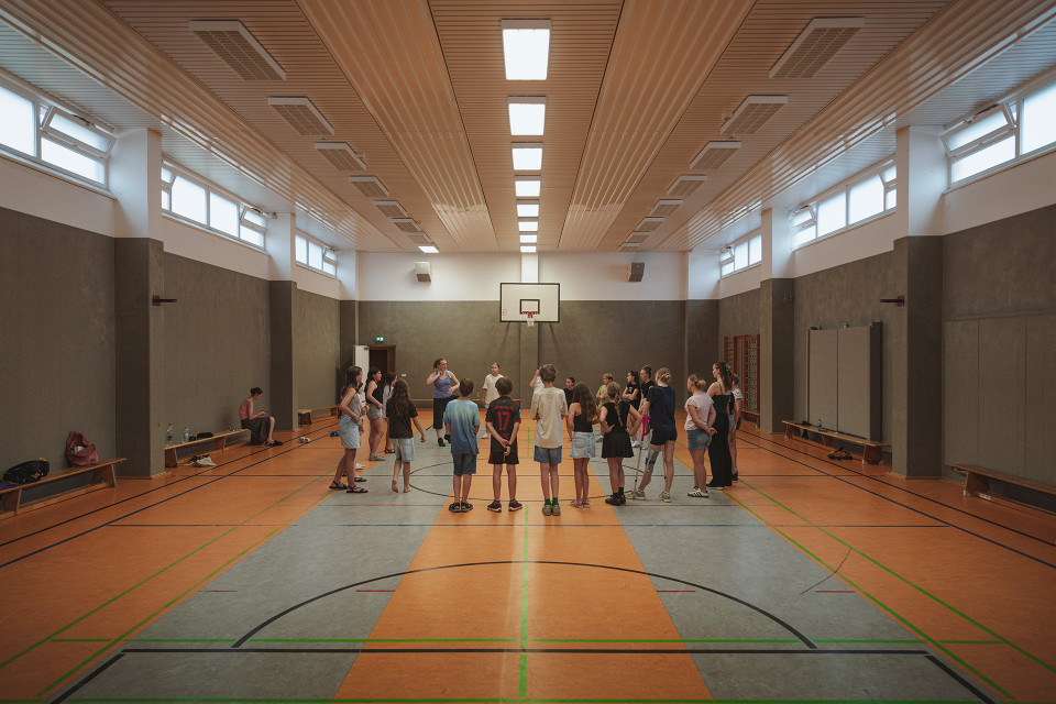 In einer großen Sporthalle steht eine Gruppe von Kindern und Jugendlichen im Kreis und hört einer Betreuerin zu. An den Wänden sind Bänke, hohe Fenster und ein Basketballkorb zu sehen.