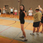 In einer Sporthalle stehen mehrere Kinder und Jugendliche verteilt auf dem Spielfeld, einige barfuß oder in Sportsachen. Im Vordergrund posiert ein Mädchen in schwarzer Kleidung mit erhobenen Fingern und herausgestreckter Zunge, während die anderen sich unterhalten oder warten.