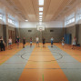 In einer großen Sporthalle stehen mehrere Kinder und Jugendliche weit verteilt entlang der Seiten und in der Mitte des Spielfelds. An der Wand hängt ein Basketballkorb, während Linien auf dem Hallenboden und hohe Fenster den Raum prägen.