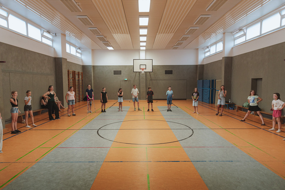 In einer großen Sporthalle stehen mehrere Kinder und Jugendliche weit verteilt entlang der Seiten und in der Mitte des Spielfelds. An der Wand hängt ein Basketballkorb, während Linien auf dem Hallenboden und hohe Fenster den Raum prägen.